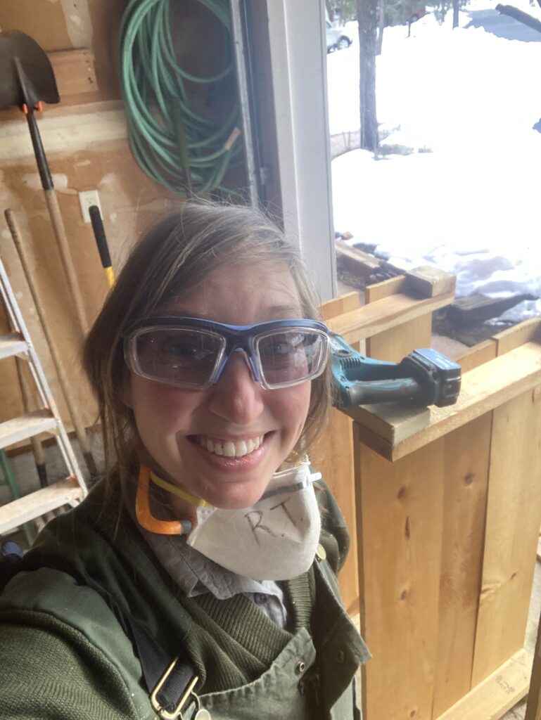 Woman in green with eye protection and ear protection smiles in front of shelves she's breaking down with a Sawz-all and hammer.