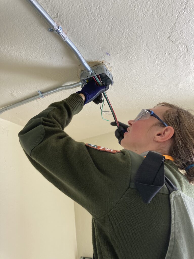 Woman in green tightens screw on electrical outlet on the ceiling.